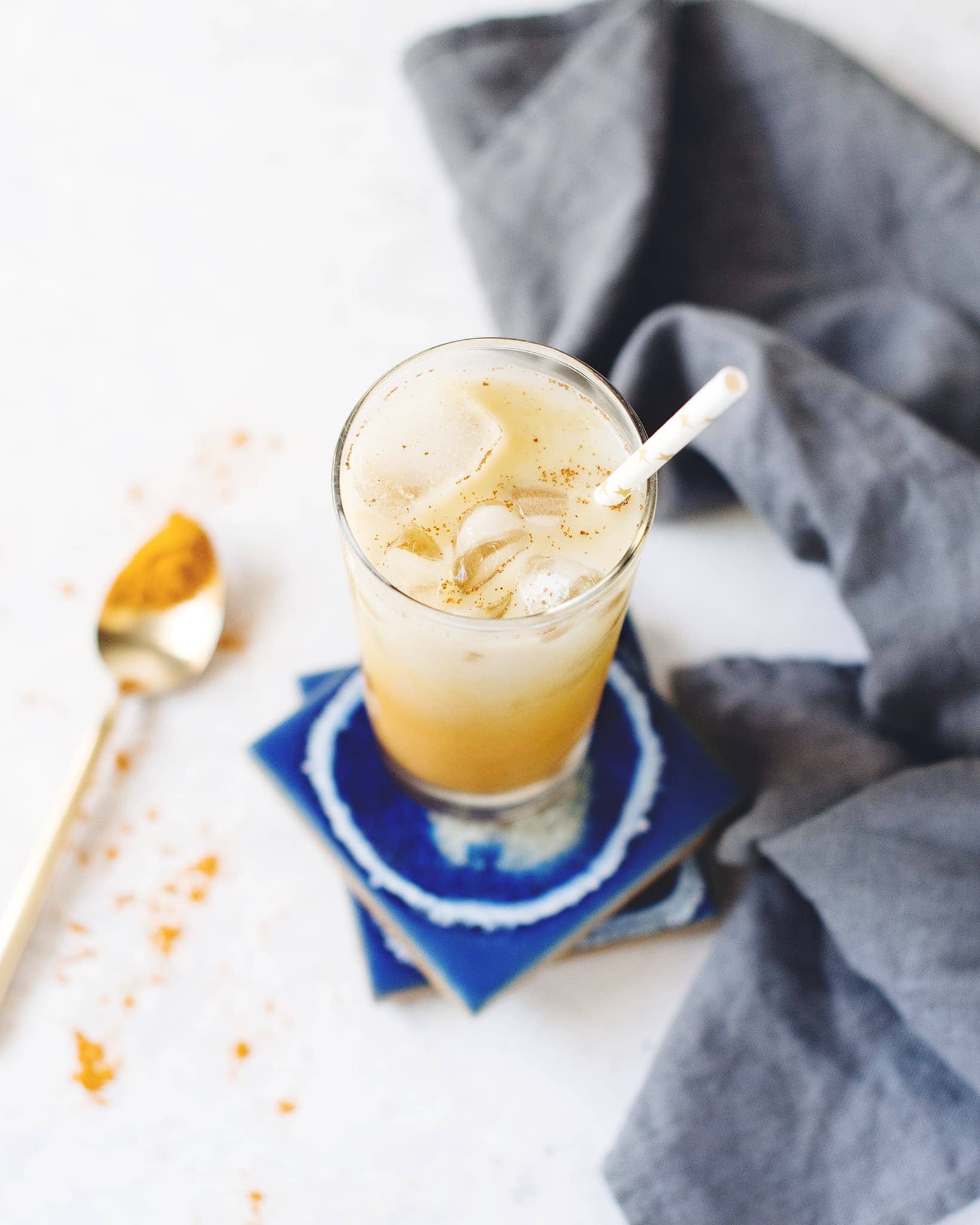 A glass of iced golden turmeric latte on blue coasters with blue linen napkin and spoon of turmeric