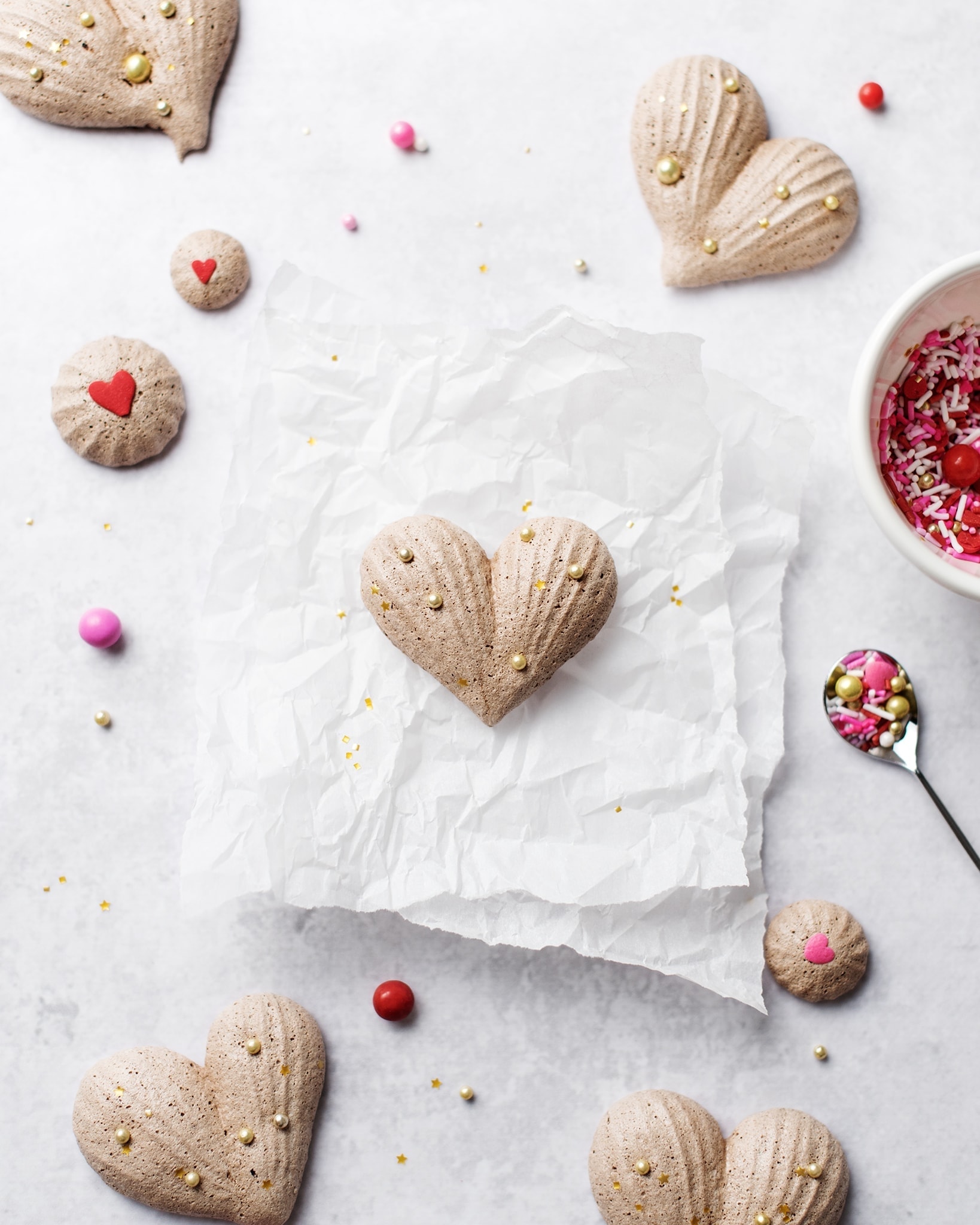 Heart-shaped chocolate meringues on parchment paper with a bowl of pink and red sprinkles