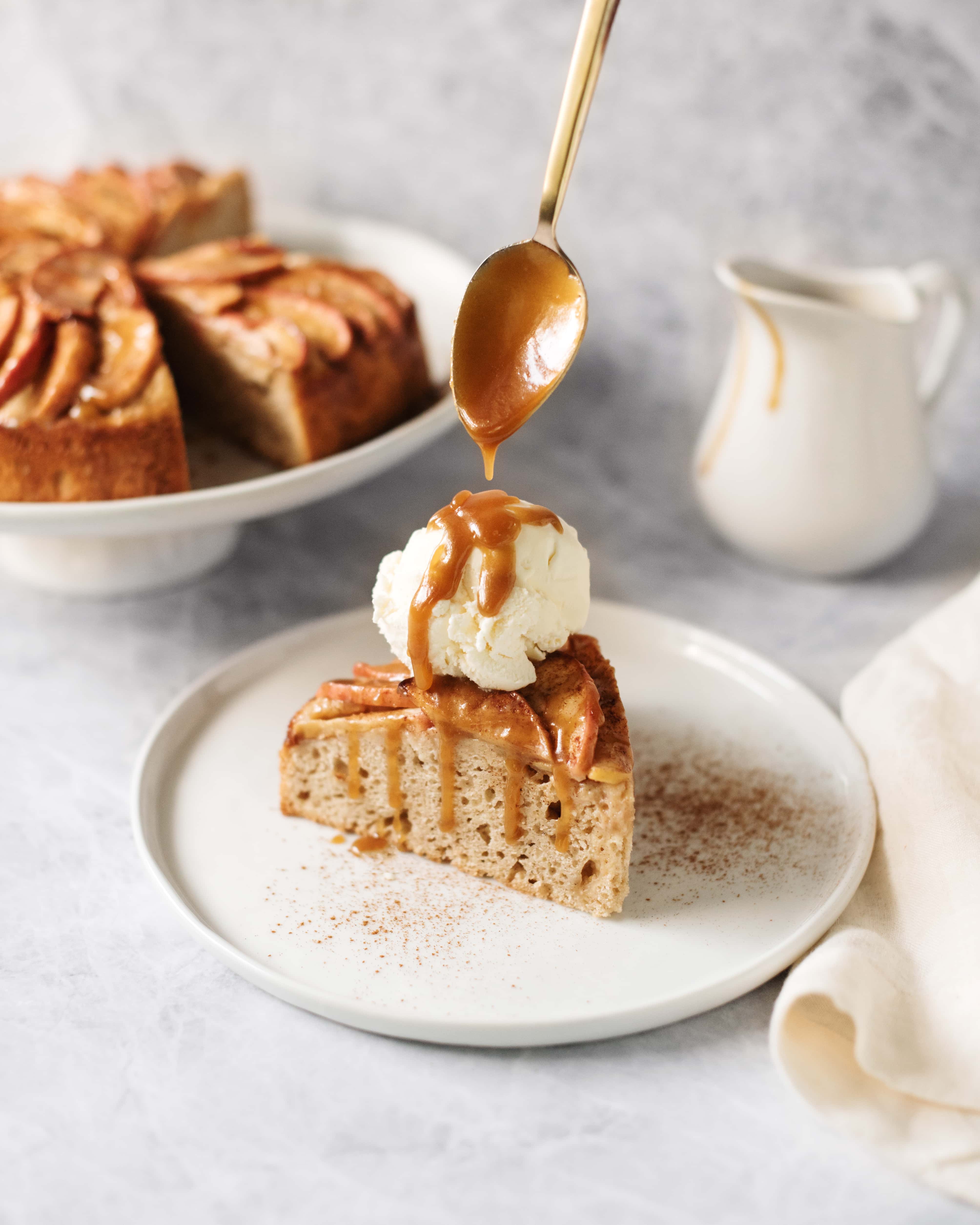 A slice of apple cake topped with a scoop of vanilla ice cream and drizzled with salted maple caramel from a spoon