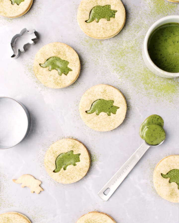 Matcha linzer cookies with triceratops cutouts on a grey background and sprinkled with matcha powder