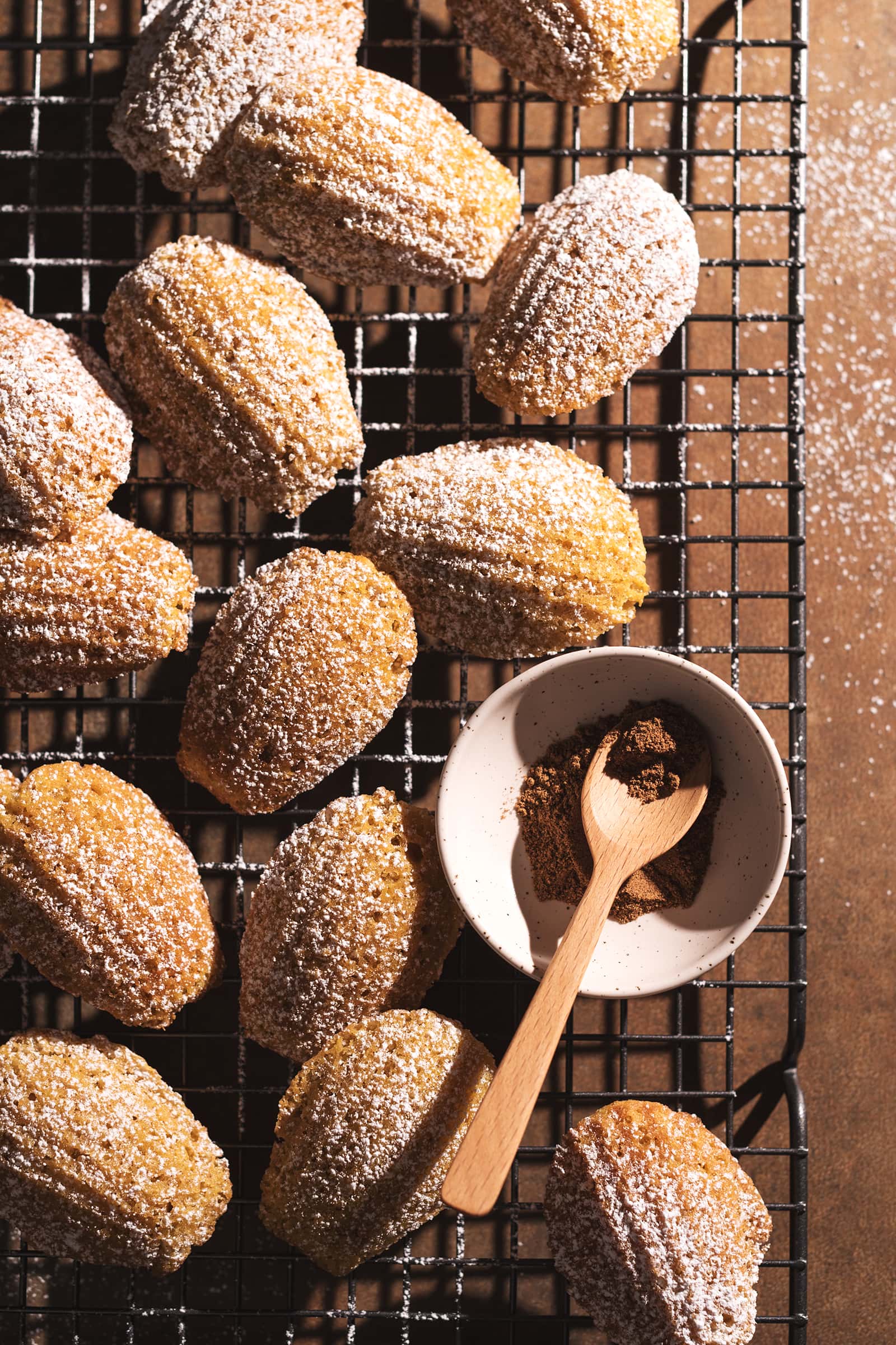 Pumpkin madeleines scattered on a wire rack with a small bowl of pumpkin pie spice.