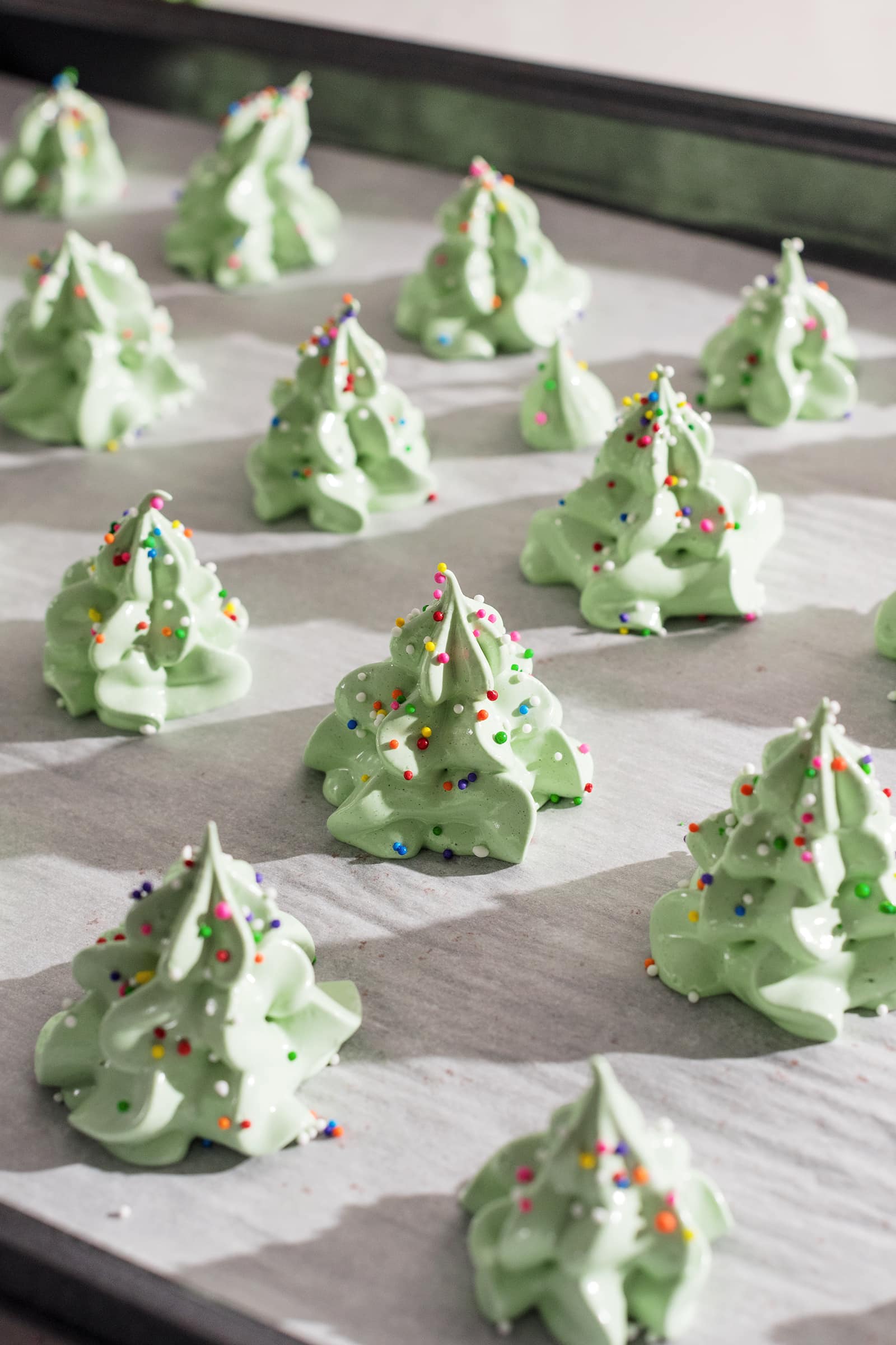 Meringue christmas trees covered in rainbow sprinkles on a baking sheet.