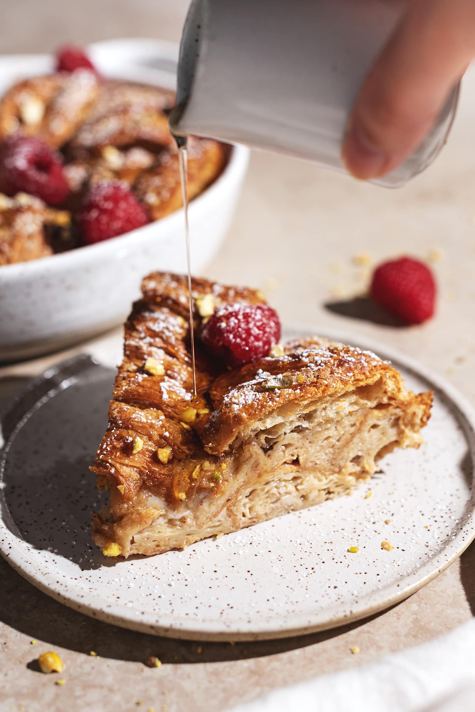 Hand pouring maple syrup onto a slice of croissant bread pudding.