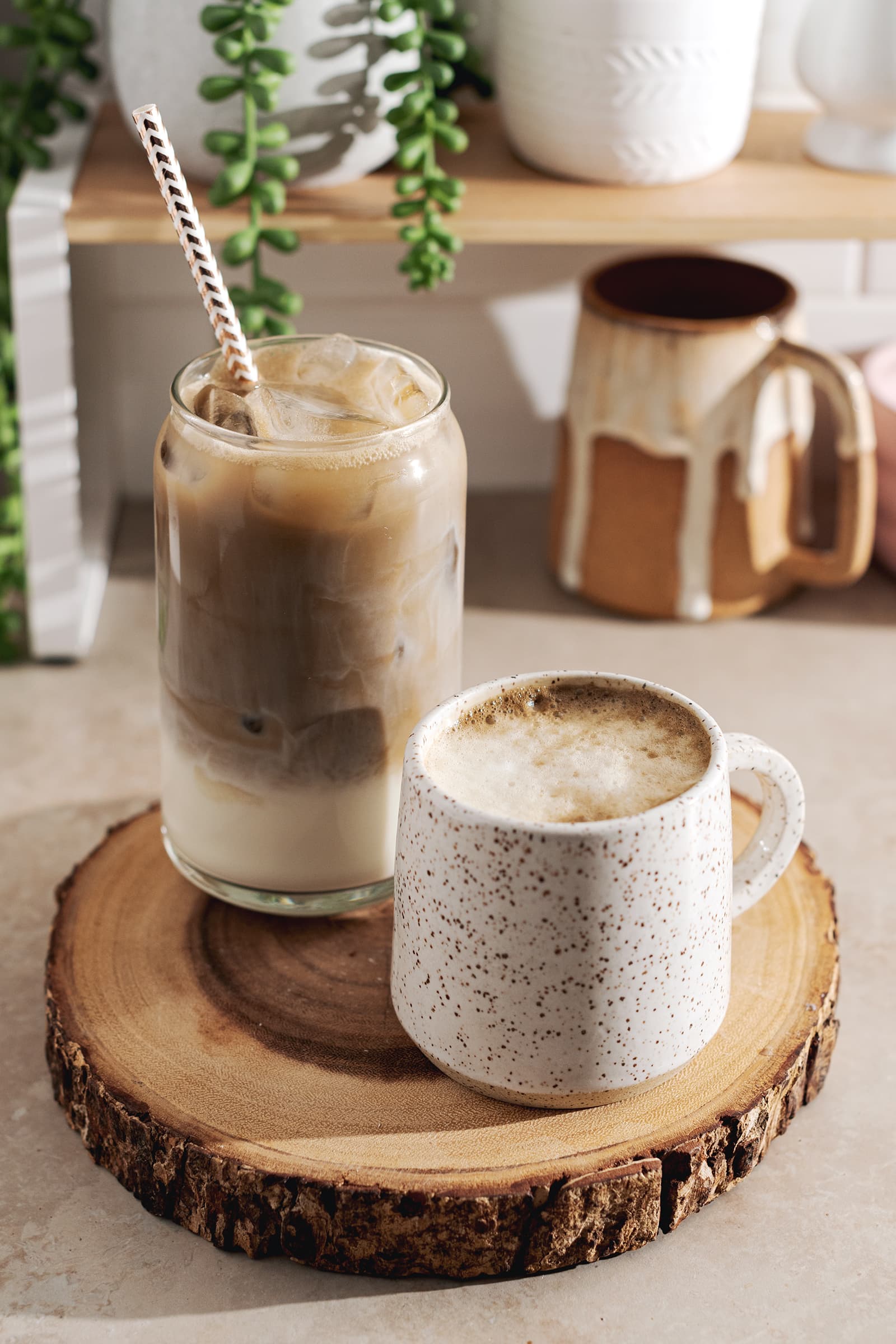 A glass of iced hojicha latte and a mug of hot hojicha latte sitting on wood platter.