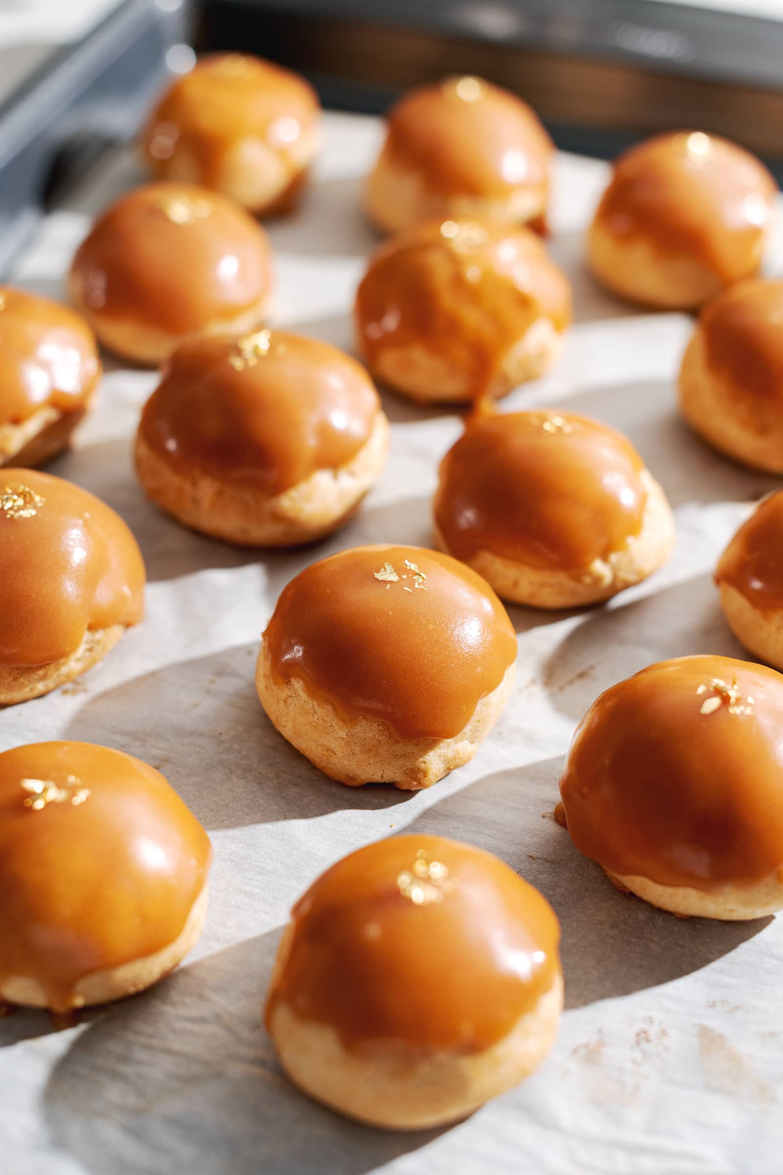 Profiteroles covered with salted caramel and gold flakes lined up parchment paper.