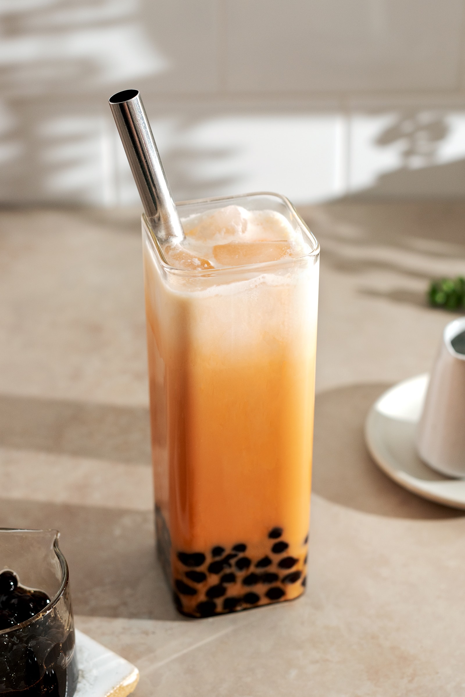 A glass of thai milk tea with boba pearls at the bottom and a layer of cream on top with a metal straw.