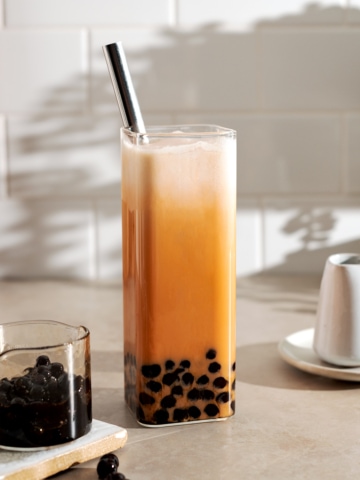 Thai milk tea boba in a rectangular glass with metal straw and small serving containers on either side.