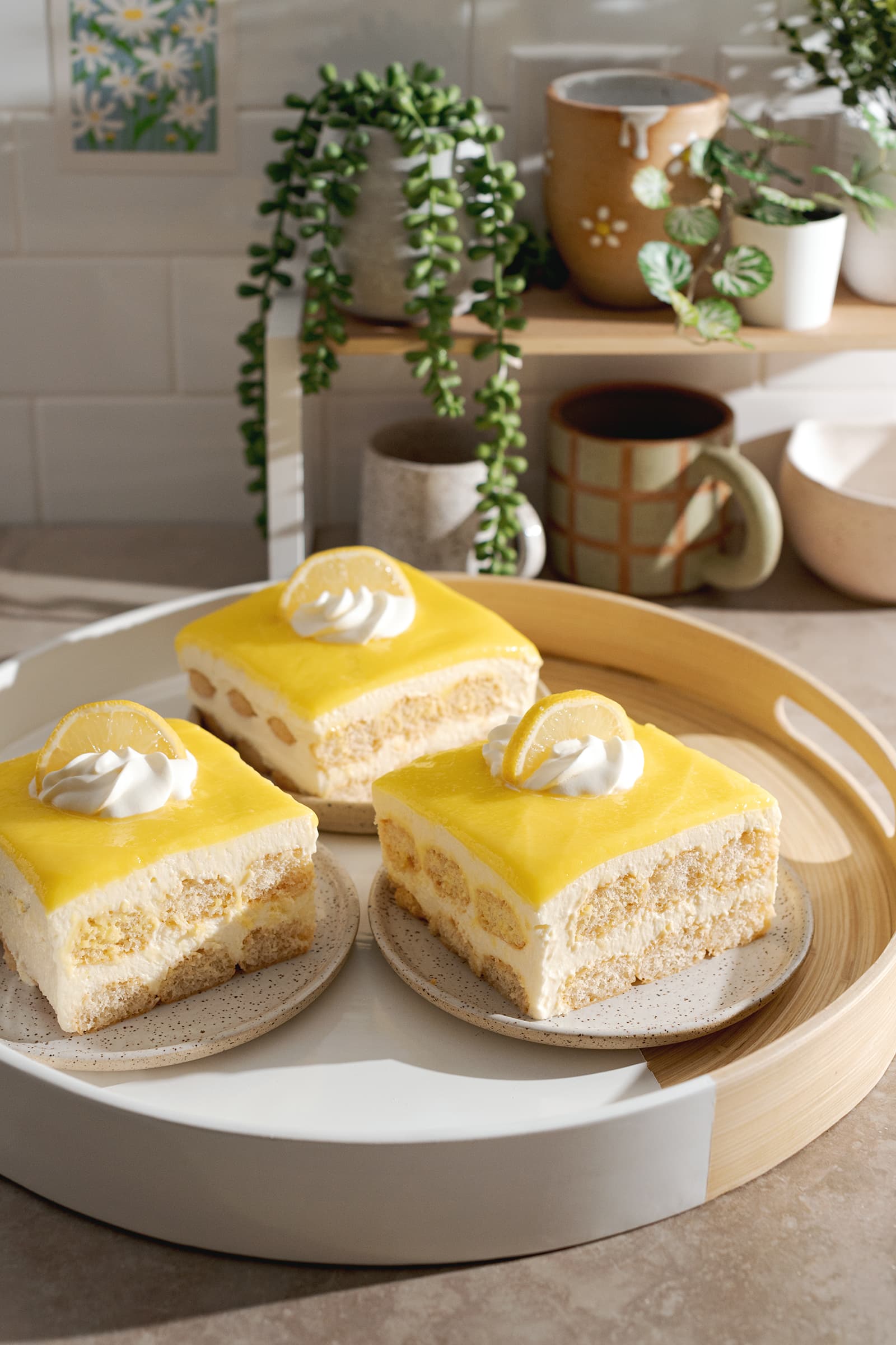 Three slices of limoncello tiramisu on a round platter in front of a shelf with plants and mugs.