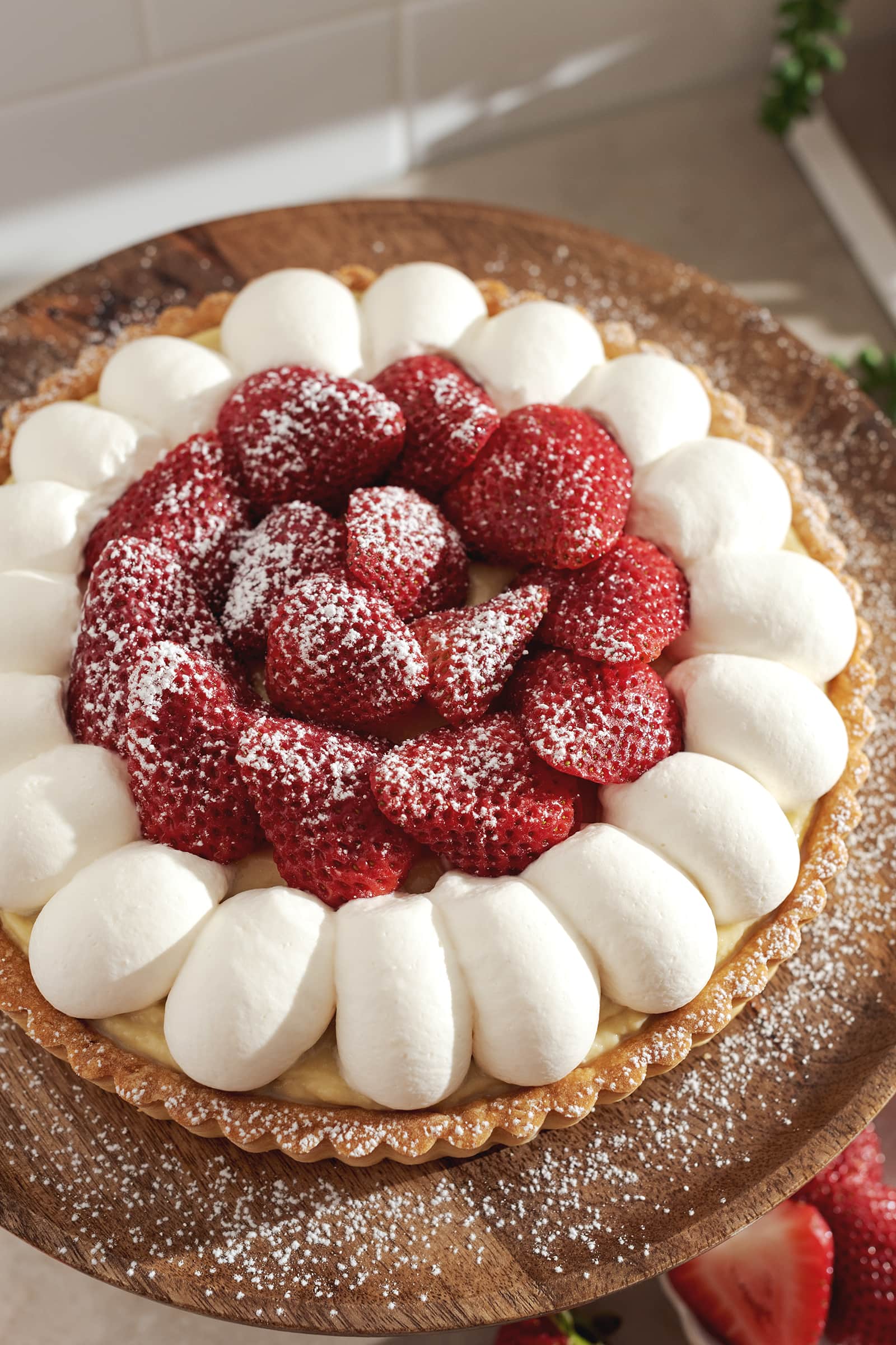 Top down view of a tart topped with whipped cream and strawberries and dusted with powdered sugar on a wooden plate.