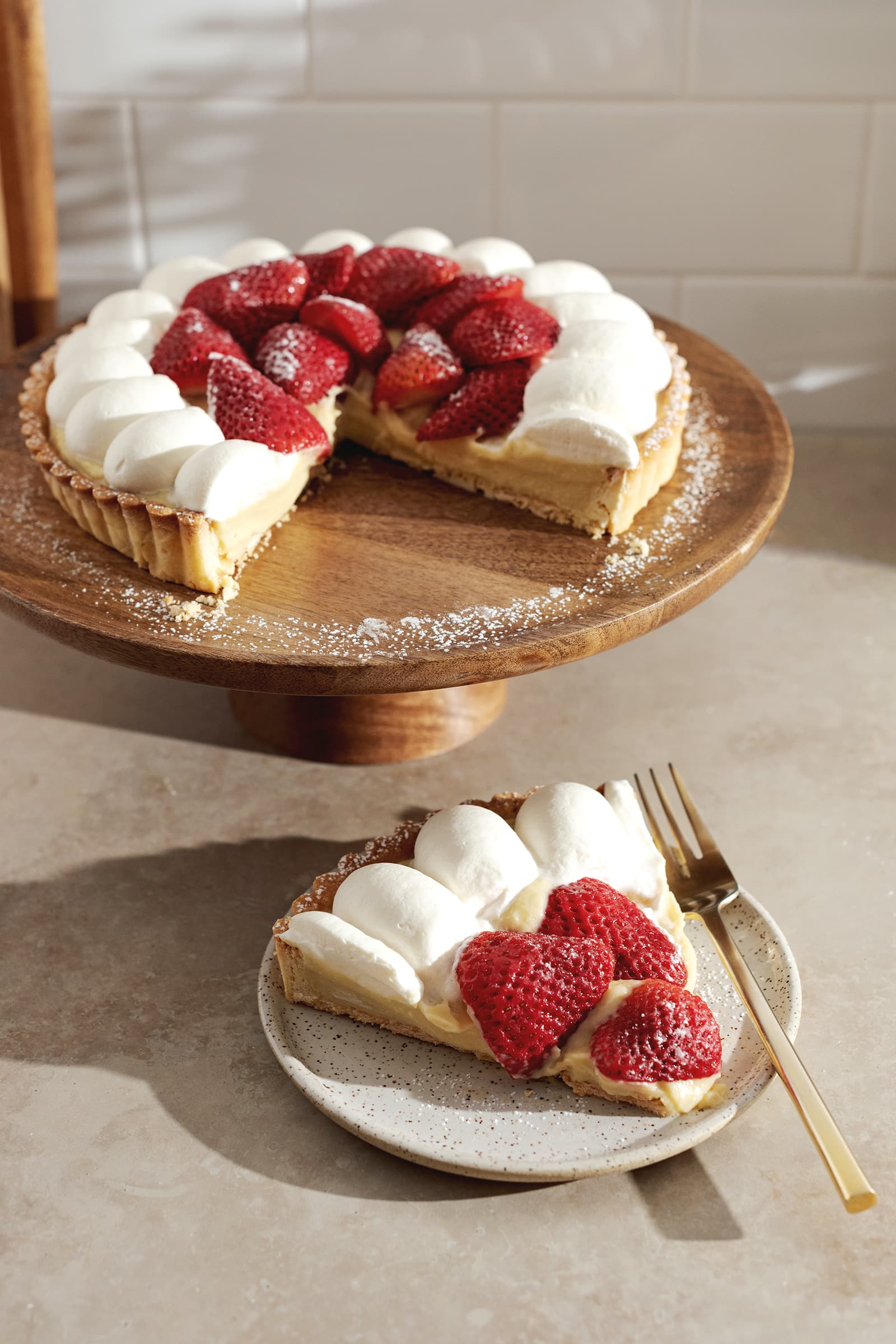 A slice of strawberry custard tart on a plate in front of the rest of the tart on a cake stand.