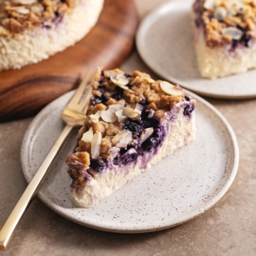 A slice of blueberry crumble cheesecake on a plate with a fork showing the layer of blueberries inside.