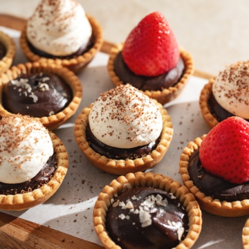 Mini chocolate tarts topped with salt flakes, whipped cream, and strawberries lined up on a wooden platter.