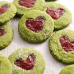 Matcha thumbprint cookies leaning against other cookies with strawberry jam in the shape of a heart.