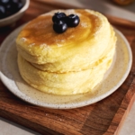A stack of two Japanese souffle pancakes with blueberries on top on a plate and wooden tray.