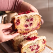 Hands holding up a slice of white chocolate raspberry loaf cake above the other slices.