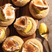 Several lemon meringue cookies scattered on a wooden board next to a spoonful of lemon curd.
