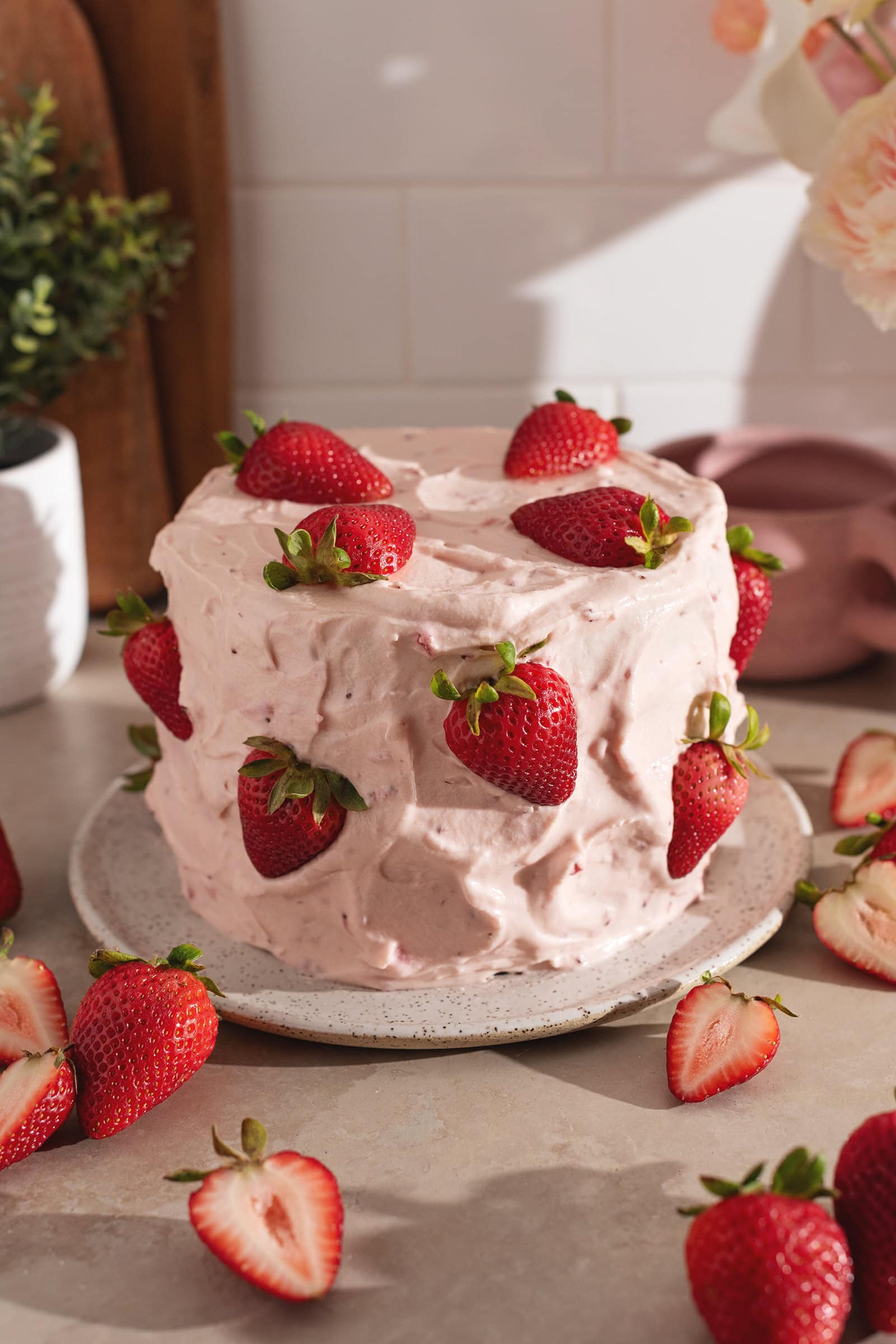A cake covered in strawberry frosting and fresh strawberries on a plate surrounded by more strawberries.