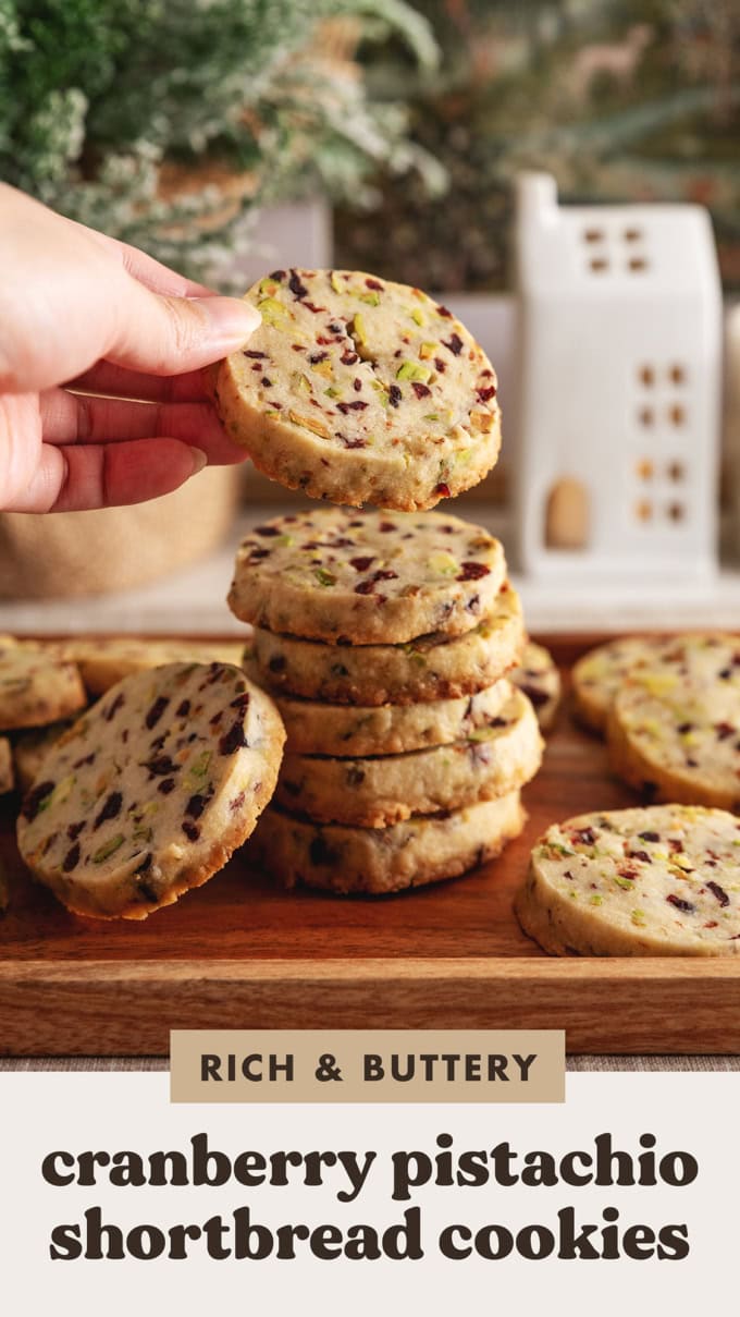 A hand holding a cranberry pistachio shortbread cookies on top of a stack of cookies.