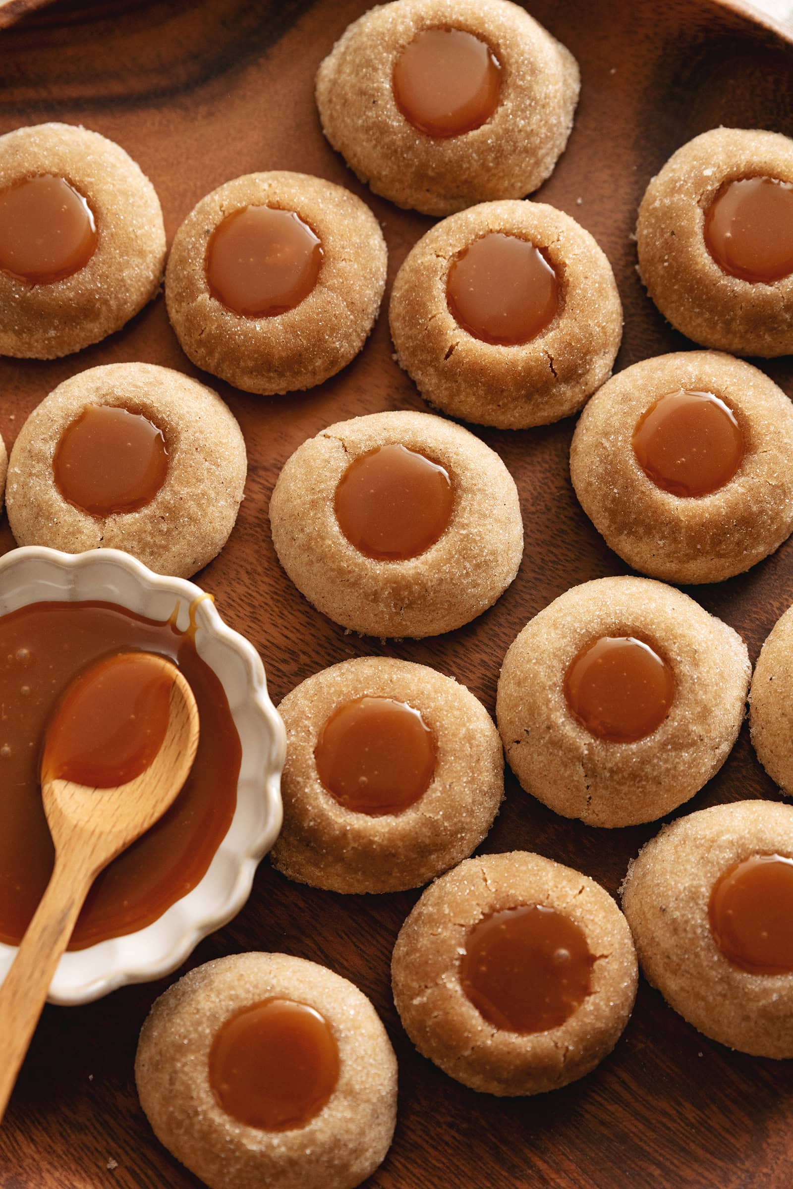 Several gingerbread thumbprint cookies scattered on a wood tray with a dish of caramel to the side.