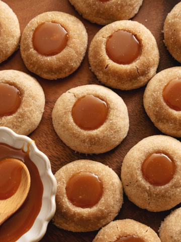 Several gingerbread thumbprint cookies scattered on a wood tray with a dish of caramel to the side.
