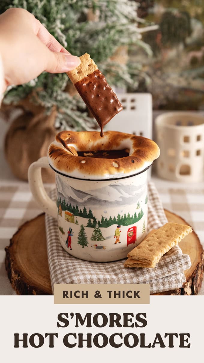 Hand dipping a graham cracker into a mug of s'mores hot chocolate with the hot chocolate dripping off the cracker.