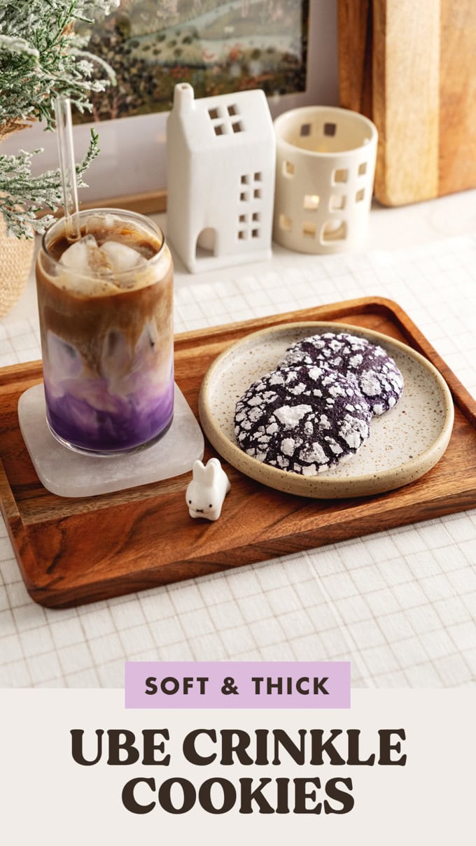 Two ube crinkle cookies on a plate with a glass of ube latte on a wooden tray.