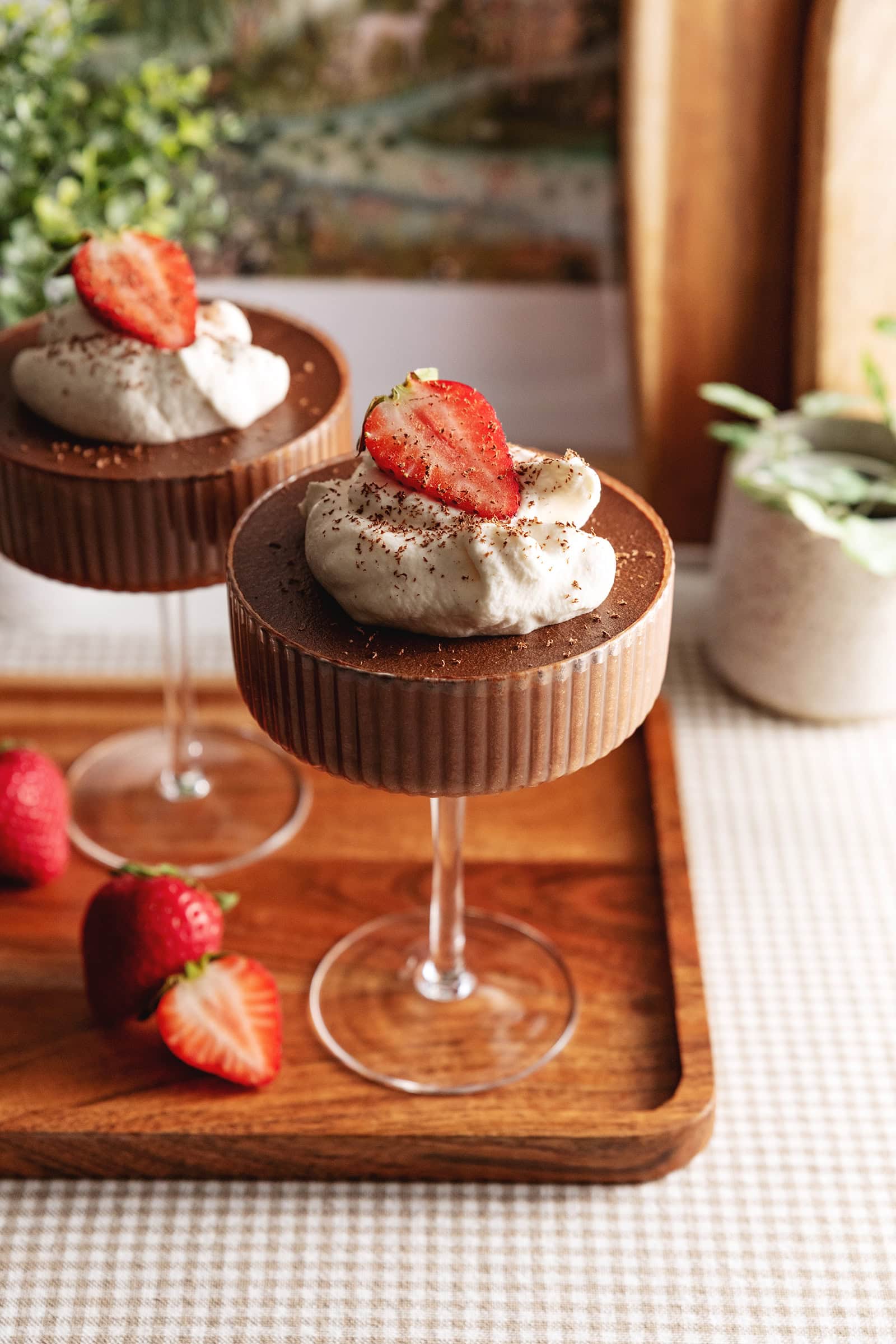 Two chocolate mousse cups in coupe glasses with whipped cream and strawberries on top on a wooden platter.