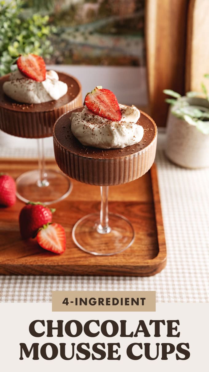 Two chocolate mousse cups in coupe glasses with whipped cream and strawberries on top on a wooden platter.